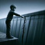 A young boy stands on a dam wall, pressing his finger into a small crack to stop water leaking through.
